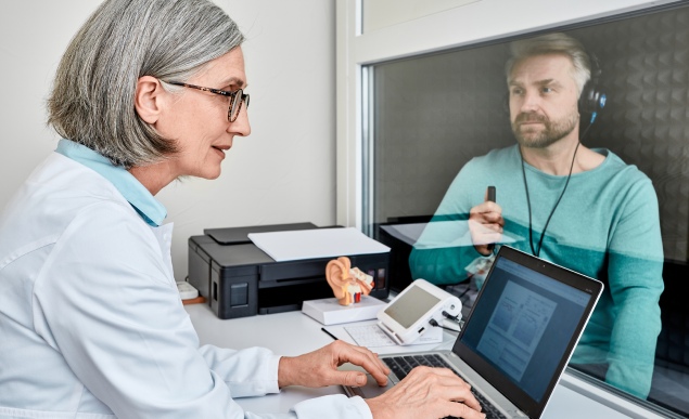 Experienced female audiologist doing hearing test to male patient in soundproof audiometric booth with audiometer in hearing clinic.